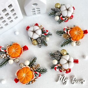 May include: Christmas-themed hair clips featuring felt cotton, orange slices, pine cones, and candy canes. The clips are adorned with faux pearls, gold glitter balls, and red ribbon accents. The clips are arranged on a white surface.