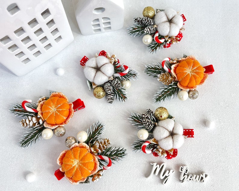 May include: Christmas-themed hair clips featuring felt cotton, orange slices, pine cones, and candy canes. The clips are adorned with faux pearls, gold glitter balls, and red ribbon accents. The clips are arranged on a white surface.