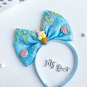 May include: A light blue headband with a large bow featuring an ice cream cone and lollipop embellishments. The bow is decorated with colorful sprinkles and the words "My Bows".