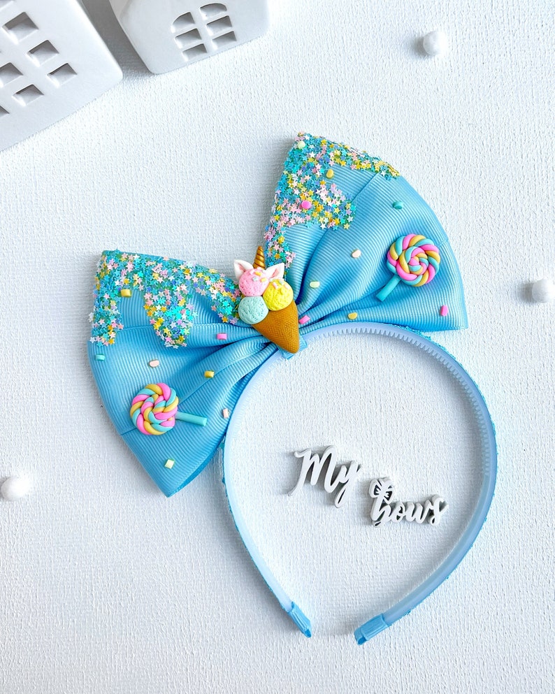 May include: A light blue headband with a large bow featuring an ice cream cone and lollipop embellishments. The bow is decorated with colorful sprinkles and the words "My Bows".