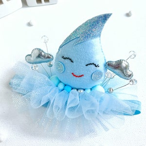 Könnte beinhalten: Ein blauer Filztropfen mit einem lächelnden Gesicht und Glitzerakzenten, umgeben von silbernen Wolken und blauem Tüll. Der Tropfen ist an einem blauen Stirnband befestigt.