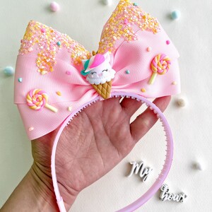 May include: A pink headband with a large bow featuring a unicorn ice cream cone design. The bow is decorated with sprinkles and lollipop accents. The headband is held in a hand.