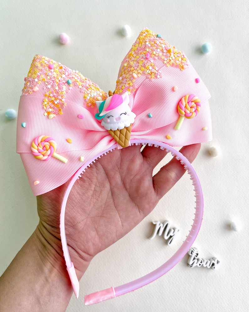 May include: A pink headband with a large bow featuring a unicorn ice cream cone design. The bow is decorated with sprinkles and lollipop accents. The headband is held in a hand.