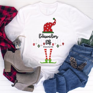 May include: White t-shirt with a graphic of an elf hat, legs, and the words "Educator Elf" and "#teacherlife". The elf hat is red with white polka dots. The legs are red and white striped, with green elf shoes.