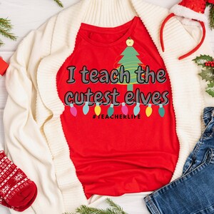 May include: A red t-shirt with the text "I teach the cutest elves" and a Christmas tree design. The shirt is part of a holiday-themed ensemble, with a white cardigan, jeans, and festive accessories.