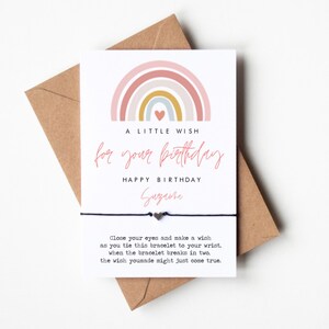 May include: A white card with a rainbow design and the text "A little wish for your birthday". The card also includes the text "Happy Birthday" and a name. The card is attached to a black string bracelet with two silver heart charms.