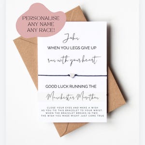 Op de afbeelding: Een witte kaart met zwarte tekst die luidt: "John, wanneer je benen het begeven, ren met je hart. Veel geluk met de Manchester Marathon." De kaart is bevestigd aan een zwart koordarmbandje met een zilveren hartbedel. De kaart bevat ook instructies voor het maken van een wens wanneer het armbandje breekt.