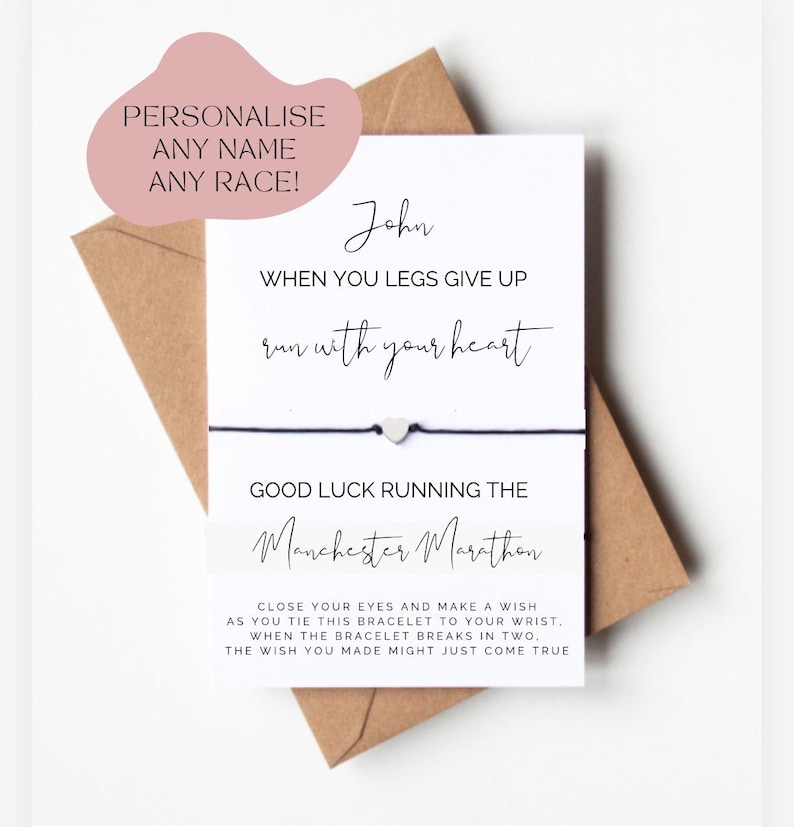 Op de afbeelding: Een witte kaart met zwarte tekst die luidt: "John, wanneer je benen het begeven, ren met je hart. Veel geluk met de Manchester Marathon." De kaart is bevestigd aan een zwart koordarmbandje met een zilveren hartbedel. De kaart bevat ook instructies voor het maken van een wens wanneer het armbandje breekt.