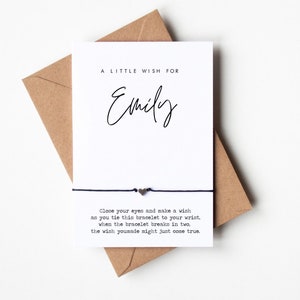 May include: A white card with black text that reads "A Little Wish For Emily". Below the text is a black string bracelet with a silver heart charm. The card reads "Close your eyes and make a wish as you tie this bracelet to your wrist, when the bracelet breaks in two, the wish you made might just come true."