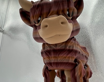 Puede incluir: Una figurita de vaca Highland escocesa con rayas marrones y blancas y una expresión amigable. La figurita tiene una superficie texturizada y está hecha de plástico.