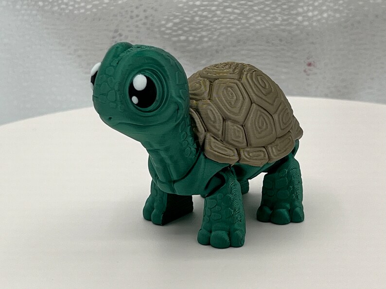 Puede incluir: Una peque&ntilde;a figura de tortuga verde y marr&oacute;n con ojos grandes. La tortuga tiene un caparaz&oacute;n detallado y est&aacute; en posici&oacute;n de caminar.