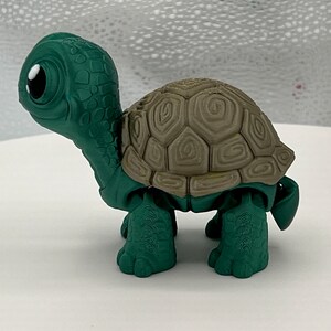 Puede incluir: Una tortuga de juguete de pl&aacute;stico verde y marr&oacute;n con un caparaz&oacute;n detallado y ojos grandes.