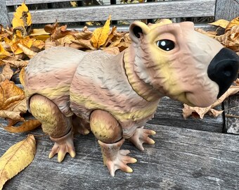 Puede incluir: Un juguete capibara marrón y amarillo con una nariz negra y ojos grandes. El juguete está hecho de plástico y tiene una superficie texturizada. El juguete está sentado en un banco de madera con hojas caídas.