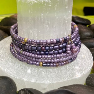 May include: A multi-strand bracelet made of purple and gold beads. The bracelet is on a white surface with a few smooth, dark grey stones.
