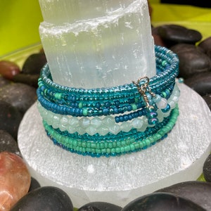 May include: A stack of beaded bracelets in shades of teal, turquoise, and seafoam green. The bracelets are wrapped around a white, rectangular crystal. The bracelets are accented with a silver charm. The bracelets are displayed on a bed of black stones.
