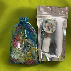 May include: A small, clear bag filled with white and blue crystals. Next to it is a silver pouch containing a small, white candle with colorful crystals embedded in the wax, a small bottle of clear liquid, and a black stone.
