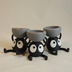 May include: Three whimsical black figurines with large white eyes and gray bowls balanced on their heads. The figures have small arms and legs, giving them a playful appearance. The background is a plain, light color.