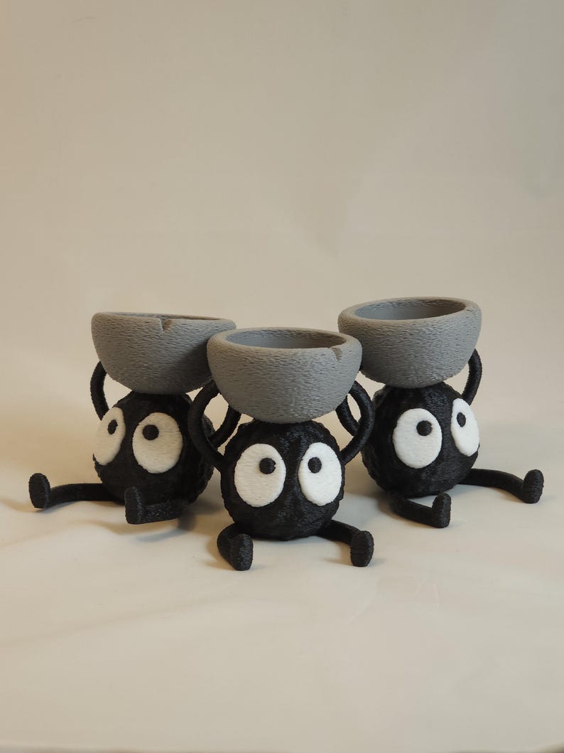 May include: Three whimsical black figurines with large white eyes and gray bowls balanced on their heads. The figures have small arms and legs, giving them a playful appearance. The background is a plain, light color.