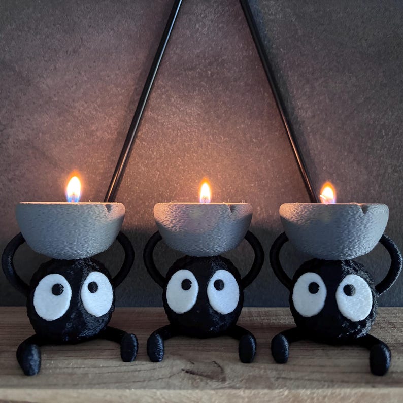 May include: Three whimsical black candle holders, each with a gray bowl holding a lit candle. The holders have large white-eyed faces and small arms and legs, creating a playful, decorative accent. The flames cast a warm glow.
