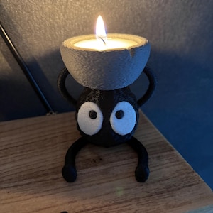 May include: A whimsical black candle holder with a cartoonish design, featuring large white eyes and a small, round candle. The candle holder is perched on a wooden surface, with a lit candle in the center.