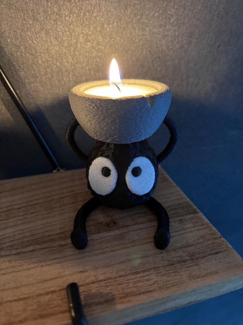 May include: A whimsical black candle holder with a cartoonish design, featuring large white eyes and a small, round candle. The candle holder is perched on a wooden surface, with a lit candle in the center.