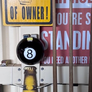 May include: A black eight ball with a white number eight, a yellow bottle opener, and a yellow sign that says "OF OWNER!". The image also includes pool cues and a red sign with white text.