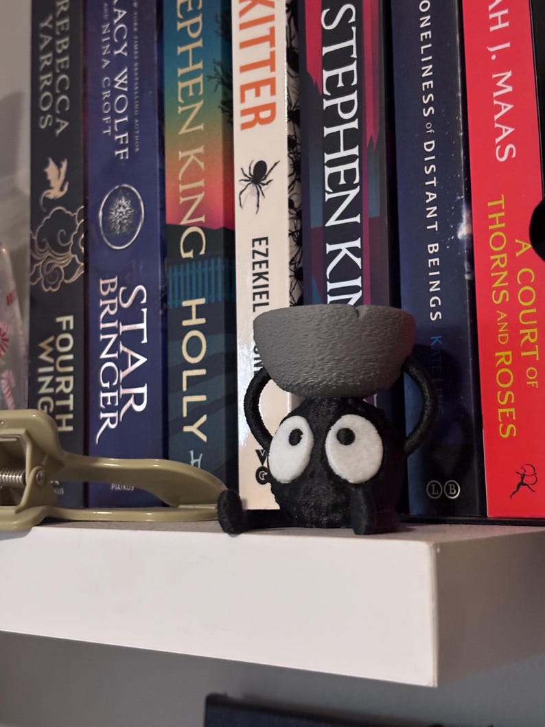 May include: A whimsical, black figurine with large white-rimmed eyes and a small bowl on its head. The figurine is perched on a white shelf in front of a row of books. The books have titles like "A Court of Thorns and Roses" and "Star Bringer".