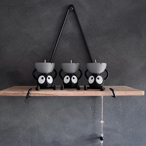 May include: Three black candle holders with large white eyes and gray candle cups sit on a wooden shelf. A black metal triangle hangs above the shelf. The shelf is mounted on a gray wall.