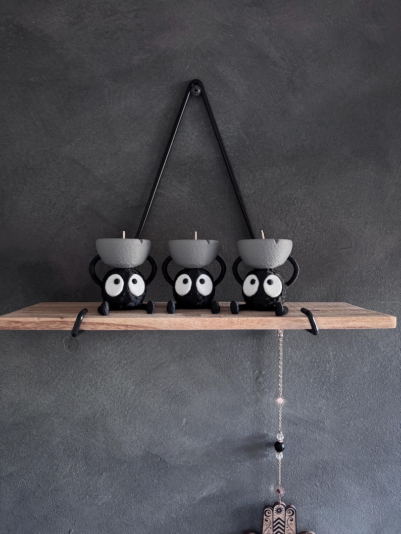May include: Three black candle holders with large white eyes and gray candle cups sit on a wooden shelf. A black metal triangle hangs above the shelf. The shelf is mounted on a gray wall.