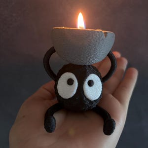 May include: A black, cartoon-like candle holder with large white eyes and a gray candle. The candle is lit with a bright orange flame. The holder has arms and legs and is being held in a hand.