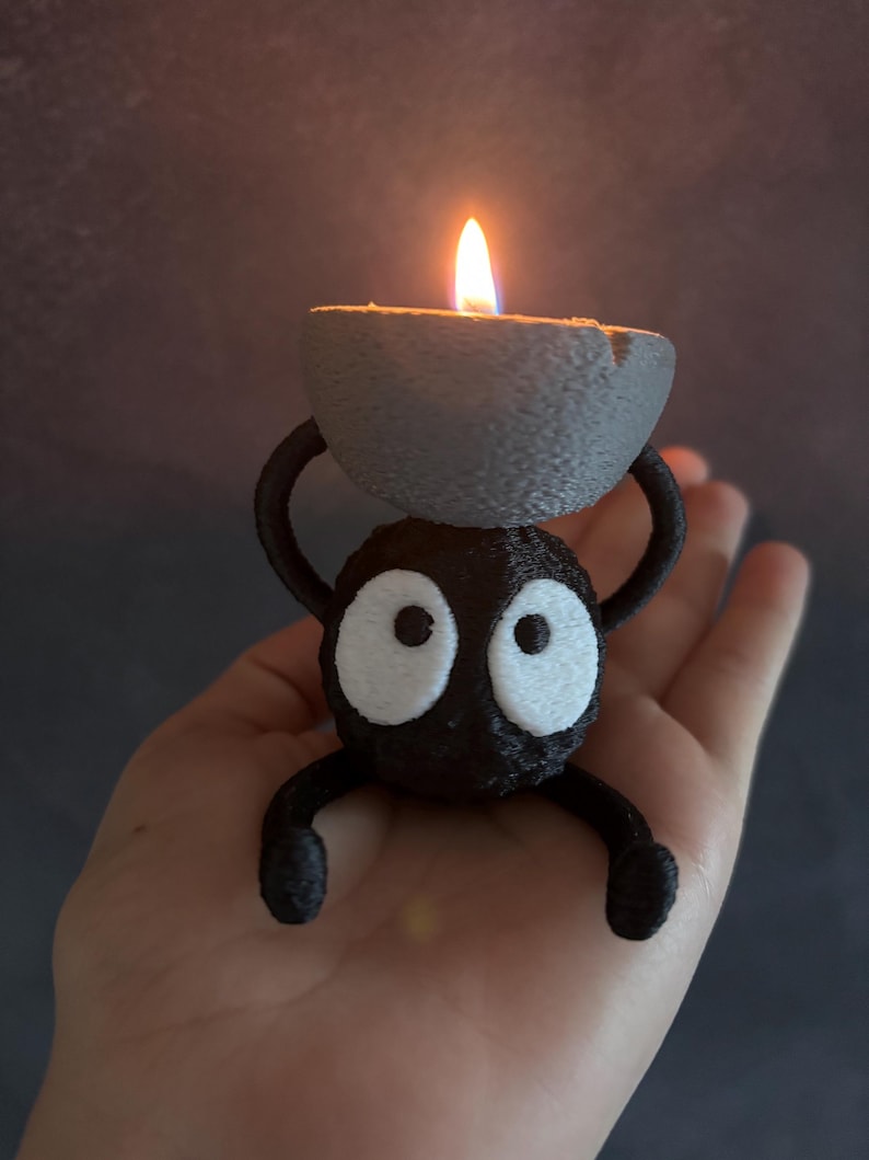May include: A black, cartoon-like candle holder with large white eyes and a gray candle. The candle is lit with a bright orange flame. The holder has arms and legs and is being held in a hand.