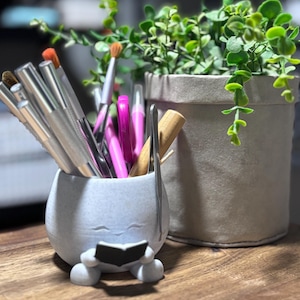 May include: A light blue ceramic holder shaped like a character reading a book, filled with makeup brushes and tools. A small potted plant sits next to the holder. The brushes have silver handles and colourful bristles.