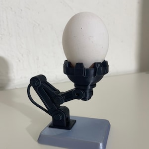 Op de afbeelding: Een wit ei dat wordt vastgehouden door een zwarte robotarm eierhouder. De houder staat op een grijze rechthoekige basis. De eierhouder heeft een mechanisch ontwerp.