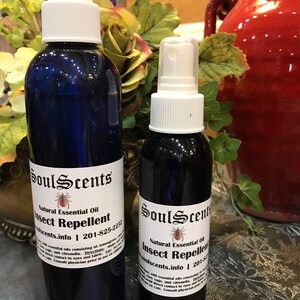 May include: Two blue glass spray bottles of SoulScents Natural Essential Oil Insect Repellent. The bottles have white labels with black text and a spider graphic. The bottles are different sizes and have white spray nozzles.