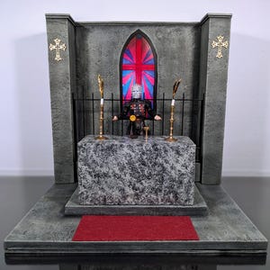 May include: A gray stone altar with a red carpet in front of it. A figure with a spiked mask stands behind a black metal gate. The figure is holding a chalice and a golden orb. A stained glass window with a red, white, and blue design is behind the figure.