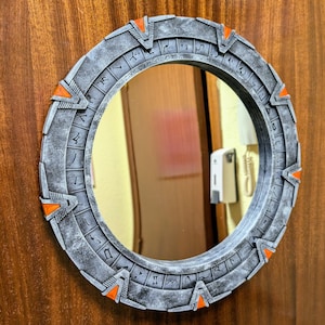 Puede incluir: Un espejo redondo de color plateado con un marco decorativo que presenta un patrón circular de símbolos y acentos naranjas. El espejo está montado en una superficie de madera.