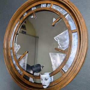 Puede incluir: Espejo redondo con marco marrón desgastado y efecto de vidrio roto. El espejo presenta una máscara blanca con una cara triste adjunta en la parte inferior. El diseño crea una estética artística única.