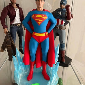 Puede incluir: Figura de acción de Superman con una capa roja y un traje azul de pie sobre una base de hielo azul y blanco. La figura tiene un cinturón rojo y el logotipo de Superman en el pecho. Dos figuras de acción más se pueden ver en el fondo.