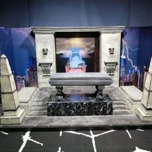 May include: A miniature diorama depicting a stone temple with a cityscape backdrop. The temple features a stone table with a pyramid and a glowing portal in the background. The diorama is made of cardboard and painted in shades of gray and white.