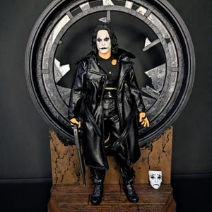 Può includere: Una action figure da collezione di un personaggio con il viso dipinto di bianco e trucco nero intorno agli occhi, che indossa un trench nero. La figura poggia su una base in legno davanti a una cornice circolare grigio scuro con dettagli argentati.