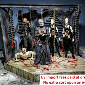 Puede incluir: Un diorama con cinco figuras de acción de Hellraiser sobre una plataforma de madera. Pinhead está en el centro sosteniendo una caja de rompecabezas, flanqueado por dos cenobitas femeninas, una figura que gatea y otra vestida de negro, unidas por cadenas.