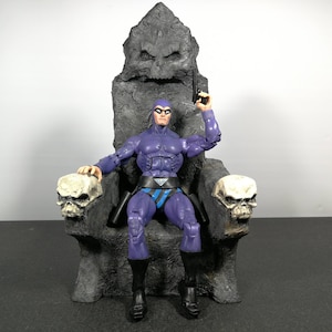 Può includere: Una action figure di un supereroe, vestito di viola e nero, seduto su un trono di pietra con braccioli a forma di teschio. La figura tiene una pistola nella mano destra.