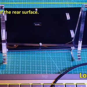 May include: A black rectangular electronic device labelled Logic Gates, shown from the rear surface. It is mounted on a clear acrylic stand and features a USB-C port. A black braided cable is connected to the device, which rests above a keyboard.