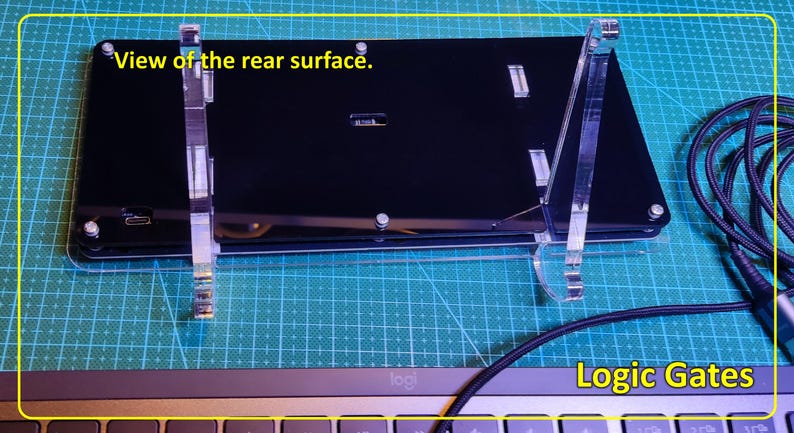 May include: A black rectangular electronic device labelled Logic Gates, shown from the rear surface. It is mounted on a clear acrylic stand and features a USB-C port. A black braided cable is connected to the device, which rests above a keyboard.