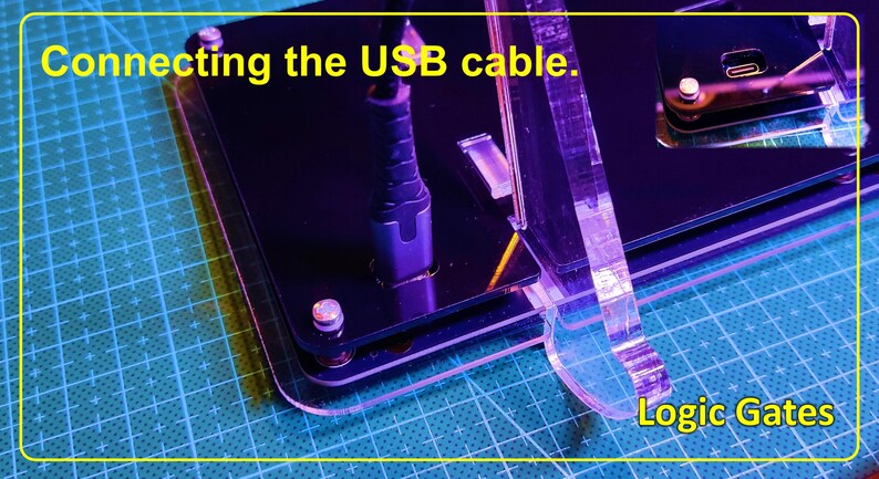 May include: A black acrylic logic gate circuit board with a USB cable connected to a port. The device features clear acrylic supports and silver hardware. The text "Connecting the USB cable." and "Logic Gates" is printed on the image.