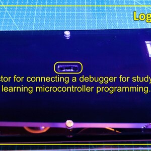 May include: A black rectangular electronic board labelled Logic Gates with a central connector port. The text on the board reads: Connector for connecting a debugger for studying and learning microcontroller programming. The board is mounted on clear supports.