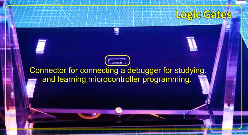 May include: A black rectangular electronic board labelled Logic Gates with a central connector port. The text on the board reads: Connector for connecting a debugger for studying and learning microcontroller programming. The board is mounted on clear supports.
