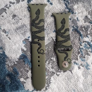 Puede incluir: Dos correas de silicona verde para Apple Watch con un diseño de serpiente negra. Las correas se muestran sobre una superficie texturizada.