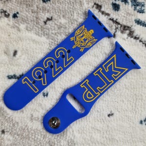 May include: A blue silicone Apple Watch band with gold lettering. One band says "1922" and the other says "DLK".