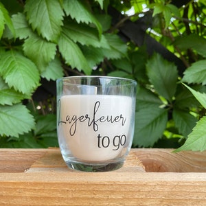Könnte beinhalten: Ein klares Glas-Kerzengefäß mit einer weißen Kerze darin. Die Kerze hat ein schwarzes Etikett mit der Aufschrift "Lagerfeuer to go".